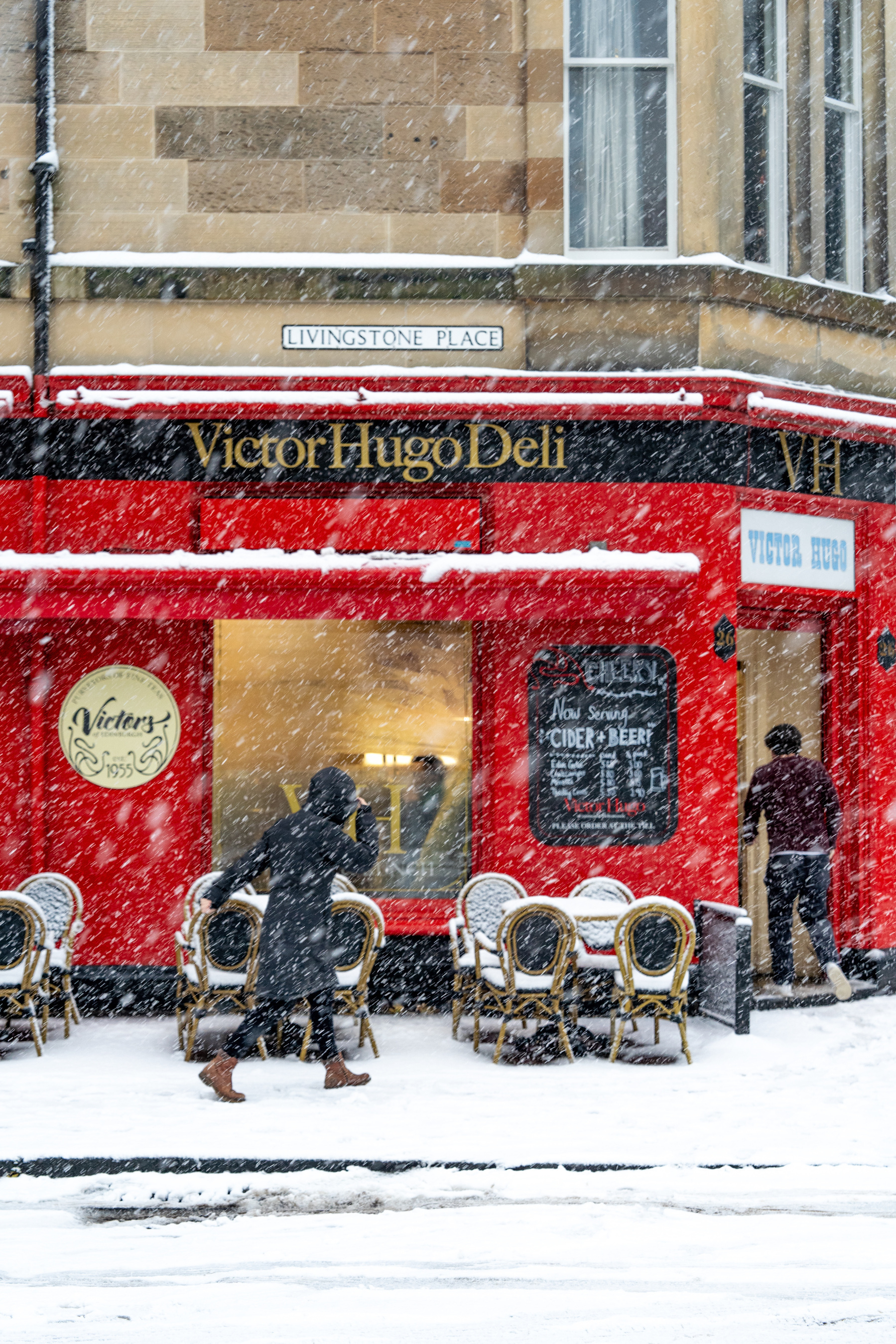 A women walks in front of Victor Hugo Deli in the snow. The cafe is bright red and welcoming. The chairs outside are covered in snow.