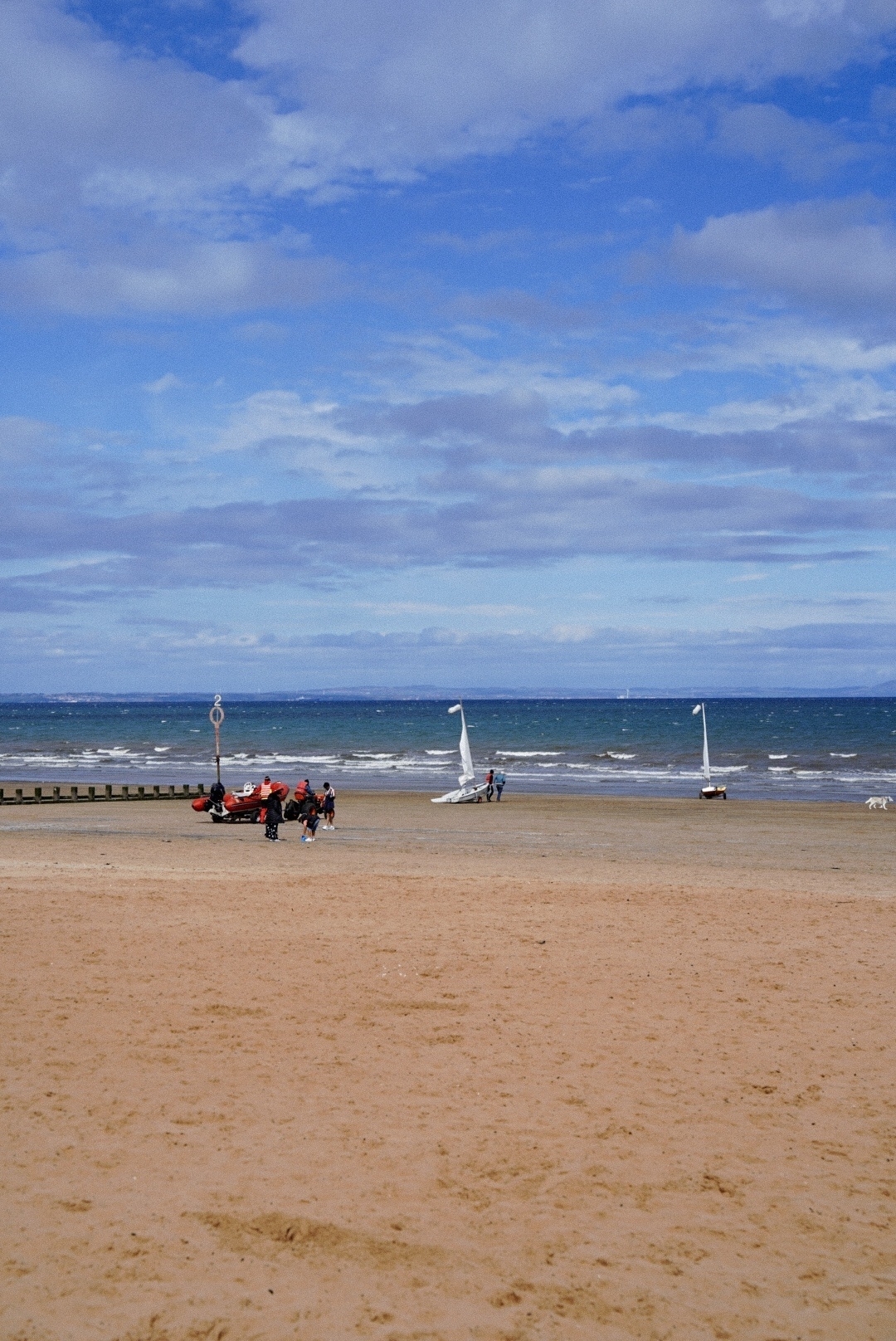Boats being wheeled across the beach towards the sea. Clouds and blue sky. 